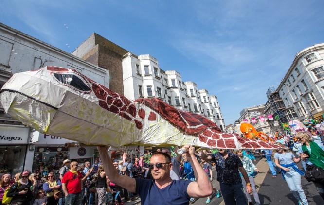 The image presents a group of people attending a parade waking through the city street. The participants are carrying large paper cut out in a shape of a turtle above their heads.