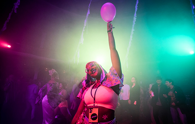A smiling person holds a balloon aloft whilst people dance behind them. st